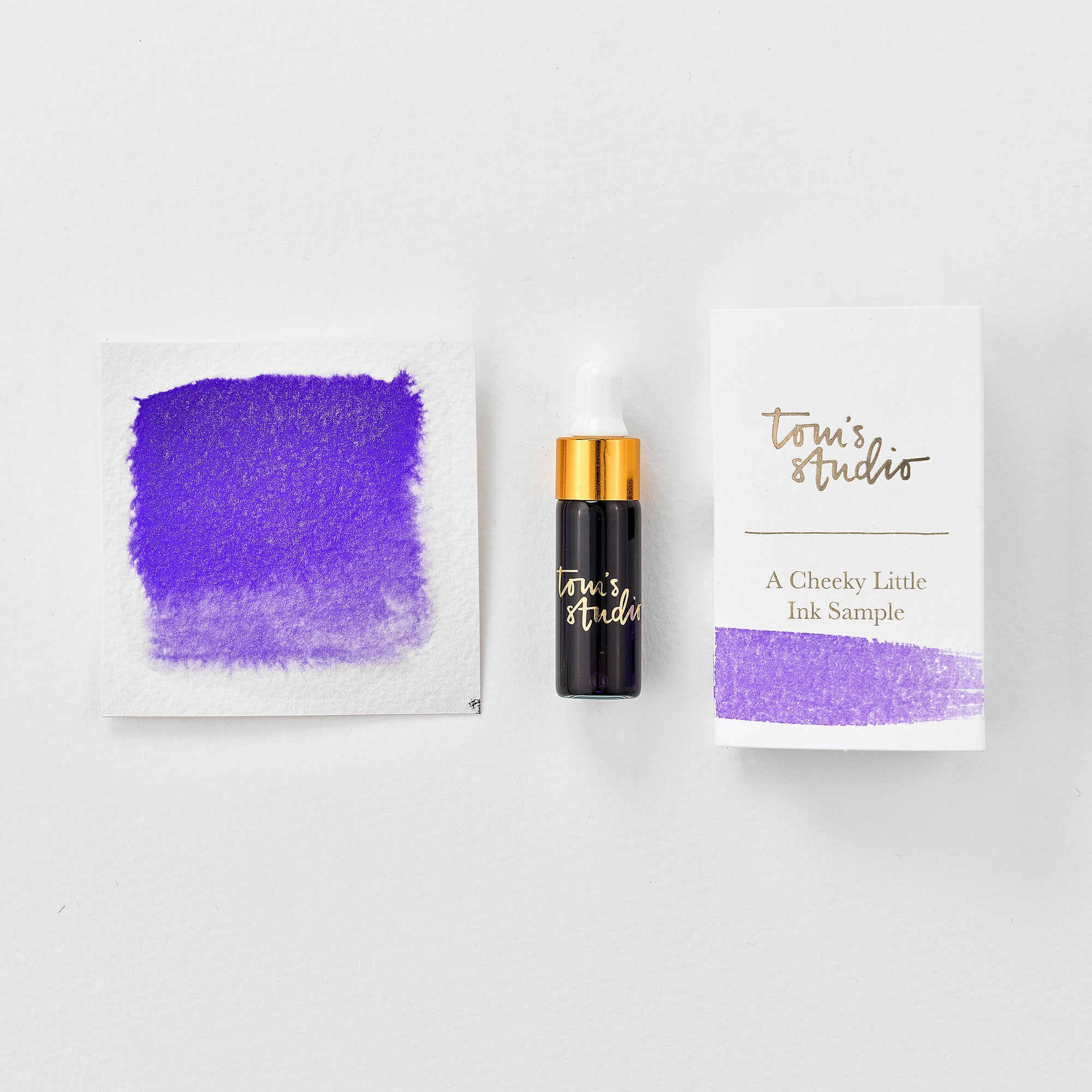 A mini ink sample bottle of Amethyst purple shimmer fountain pen ink by Tom’s Studio, displayed alongside a swatch of the ink on textured paper and its matching packaging. The bottle is small, with a gold cap and a white pipette dropper, and features 'Tom’s Studio' in gold lettering. The ink swatch shows a rich, shimmering hue with a gradient from deep to lighter tones. The packaging, a small white card, has 'Tom’s Studio' embossed in gold at the top, with a brushstroke of the ink colour at the bottom.