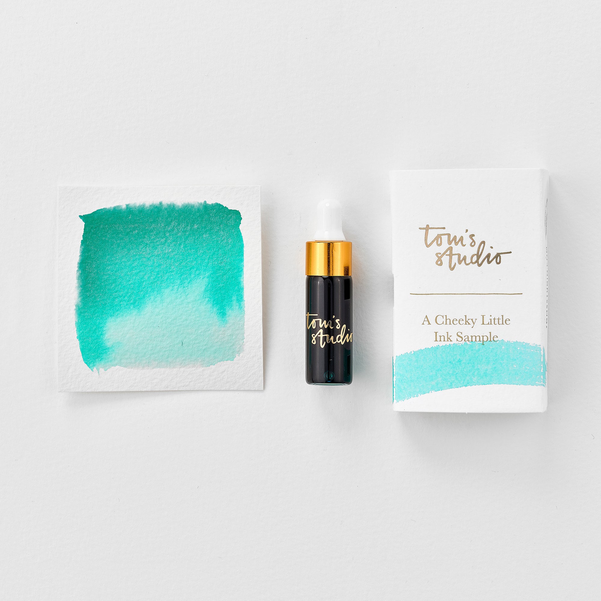 A mini ink sample bottle of Barrier Reef blue shimmer fountain pen ink by Tom’s Studio, displayed alongside a swatch of the ink on textured paper and its matching packaging. The bottle is small, with a gold cap and a white pipette dropper, and features 'Tom’s Studio' in gold lettering. The ink swatch shows a rich, shimmering hue with a gradient from deep to lighter tones. The packaging, a small white card, has 'Tom’s Studio' embossed in gold at the top, with a brushstroke of the ink colour at the bottom.