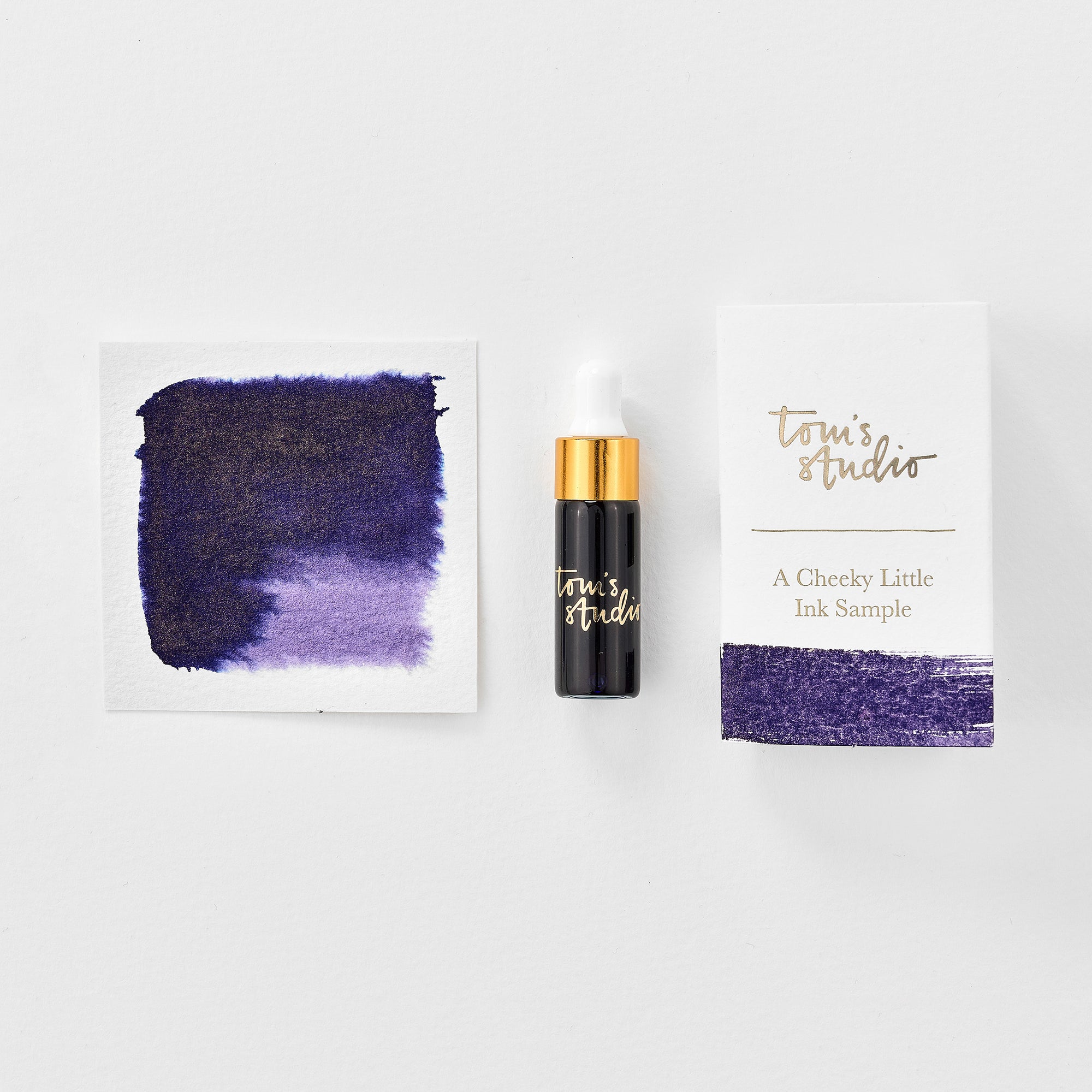 A mini ink sample bottle of Broadway purple shimmer fountain pen ink by Tom’s Studio, displayed alongside a swatch of the ink on textured paper and its matching packaging. The bottle is small, with a gold cap and a white pipette dropper, and features 'Tom’s Studio' in gold lettering. The ink swatch shows a rich, shimmering hue with a gradient from deep to lighter tones. The packaging, a small white card, has 'Tom’s Studio' embossed in gold at the top, with a brushstroke of the ink colour at the bottom.