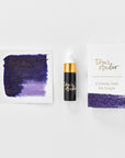 A mini ink sample bottle of Broadway purple shimmer fountain pen ink by Tom’s Studio, displayed alongside a swatch of the ink on textured paper and its matching packaging. The bottle is small, with a gold cap and a white pipette dropper, and features 'Tom’s Studio' in gold lettering. The ink swatch shows a rich, shimmering hue with a gradient from deep to lighter tones. The packaging, a small white card, has 'Tom’s Studio' embossed in gold at the top, with a brushstroke of the ink colour at the bottom.