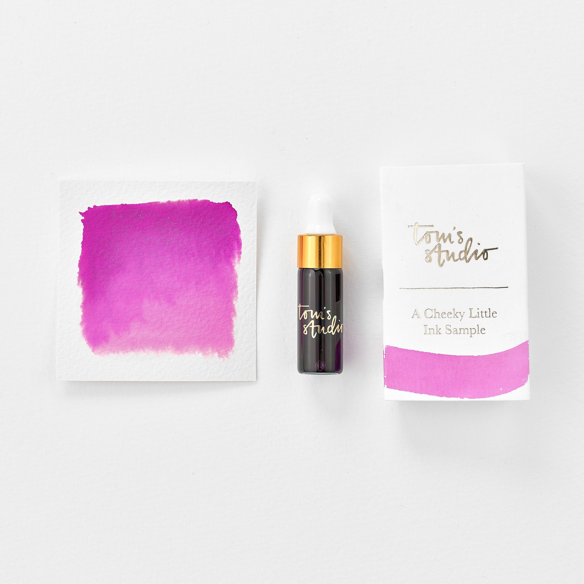 A mini ink sample bottle of Cherry Bomb pink shimmer fountain pen ink by Tom’s Studio, displayed alongside a swatch of the ink on textured paper and its matching packaging. The bottle is small, with a gold cap and a white pipette dropper, and features 'Tom’s Studio' in gold lettering. The ink swatch shows a rich, shimmering hue with a gradient from deep to lighter tones. The packaging, a small white card, has 'Tom’s Studio' embossed in gold at the top, with a brushstroke of the ink colour at the bottom.