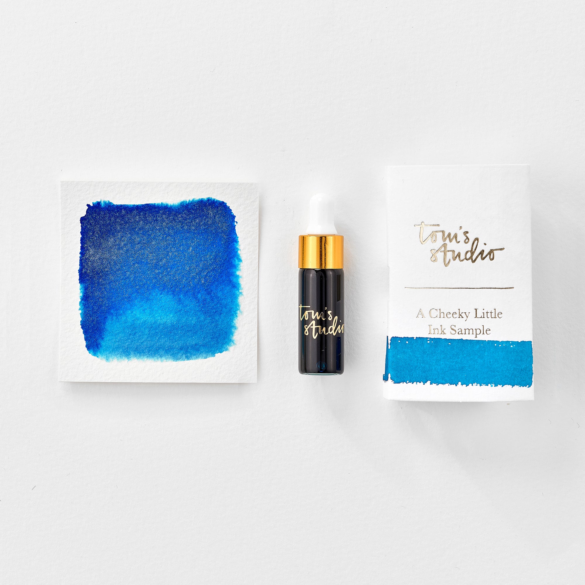 A mini ink sample bottle of Kingfisher blue shimmer fountain pen ink by Tom’s Studio, displayed alongside a swatch of the ink on textured paper and its matching packaging. The bottle is small, with a gold cap and a white pipette dropper, and features 'Tom’s Studio' in gold lettering. The ink swatch shows a rich, shimmering hue with a gradient from deep to lighter tones. The packaging, a small white card, has 'Tom’s Studio' embossed in gold at the top, with a brushstroke of the ink colour at the bottom.