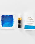 A mini ink sample bottle of Kingfisher blue shimmer fountain pen ink by Tom’s Studio, displayed alongside a swatch of the ink on textured paper and its matching packaging. The bottle is small, with a gold cap and a white pipette dropper, and features 'Tom’s Studio' in gold lettering. The ink swatch shows a rich, shimmering hue with a gradient from deep to lighter tones. The packaging, a small white card, has 'Tom’s Studio' embossed in gold at the top, with a brushstroke of the ink colour at the bottom.