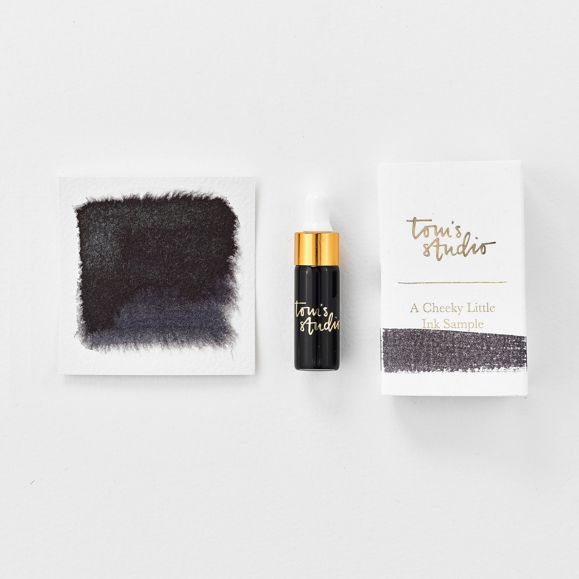 A mini ink sample bottle of Stellar black shimmer fountain pen ink by Tom’s Studio, displayed alongside a swatch of the ink on textured paper and its matching packaging. The bottle is small, with a gold cap and a white pipette dropper, and features 'Tom’s Studio' in gold lettering. The ink swatch shows a rich, shimmering hue with a gradient from deep to lighter tones. The packaging, a small white card, has 'Tom’s Studio' embossed in gold at the top, with a brushstroke of the ink colour at the bottom.