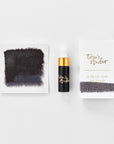 A mini ink sample bottle of Stellar black shimmer fountain pen ink by Tom’s Studio, displayed alongside a swatch of the ink on textured paper and its matching packaging. The bottle is small, with a gold cap and a white pipette dropper, and features 'Tom’s Studio' in gold lettering. The ink swatch shows a rich, shimmering hue with a gradient from deep to lighter tones. The packaging, a small white card, has 'Tom’s Studio' embossed in gold at the top, with a brushstroke of the ink colour at the bottom.