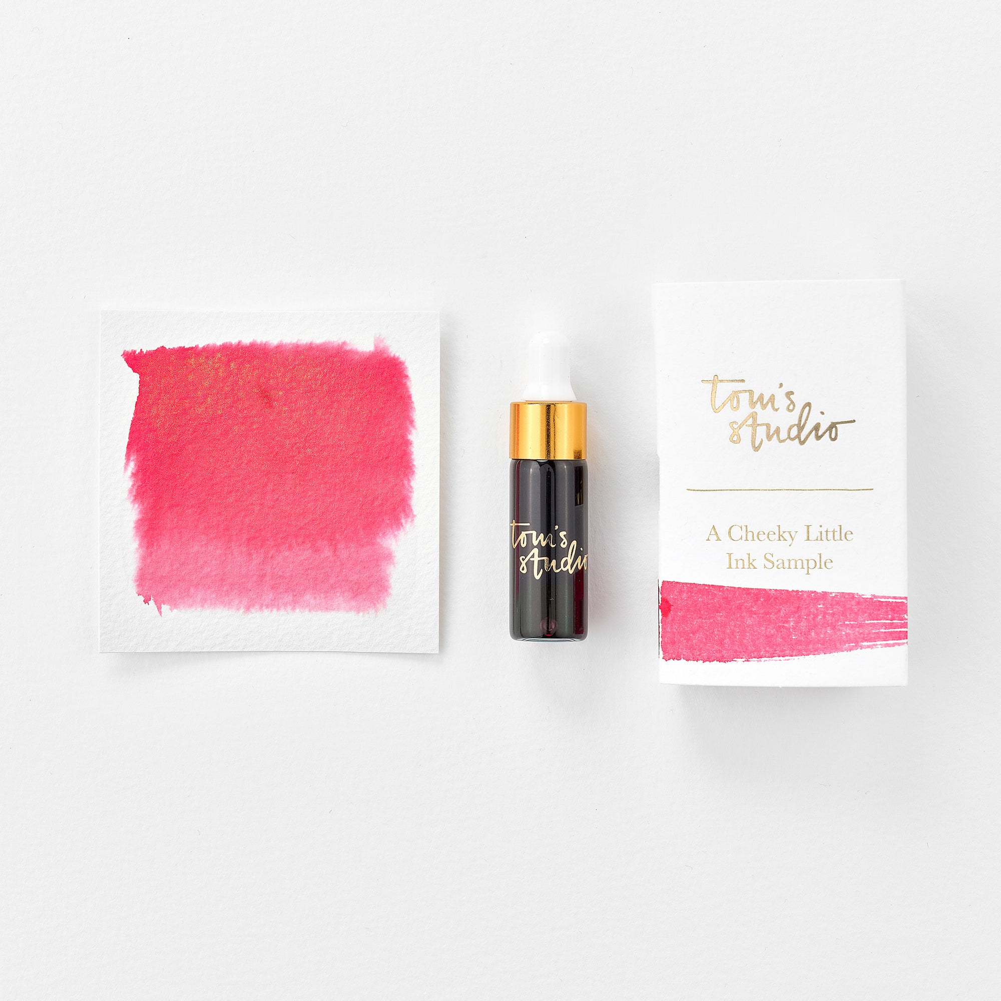 A mini ink sample bottle of Watermelon pink shimmer fountain pen ink by Tom’s Studio, displayed alongside a swatch of the ink on textured paper and its matching packaging. The bottle is small, with a gold cap and a white pipette dropper, and features 'Tom’s Studio' in gold lettering. The ink swatch shows a rich, shimmering hue with a gradient from deep to lighter tones. The packaging, a small white card, has 'Tom’s Studio' embossed in gold at the top, with a brushstroke of the ink colour at the bottom.