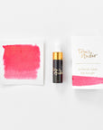 A mini ink sample bottle of Watermelon pink shimmer fountain pen ink by Tom’s Studio, displayed alongside a swatch of the ink on textured paper and its matching packaging. The bottle is small, with a gold cap and a white pipette dropper, and features 'Tom’s Studio' in gold lettering. The ink swatch shows a rich, shimmering hue with a gradient from deep to lighter tones. The packaging, a small white card, has 'Tom’s Studio' embossed in gold at the top, with a brushstroke of the ink colour at the bottom.