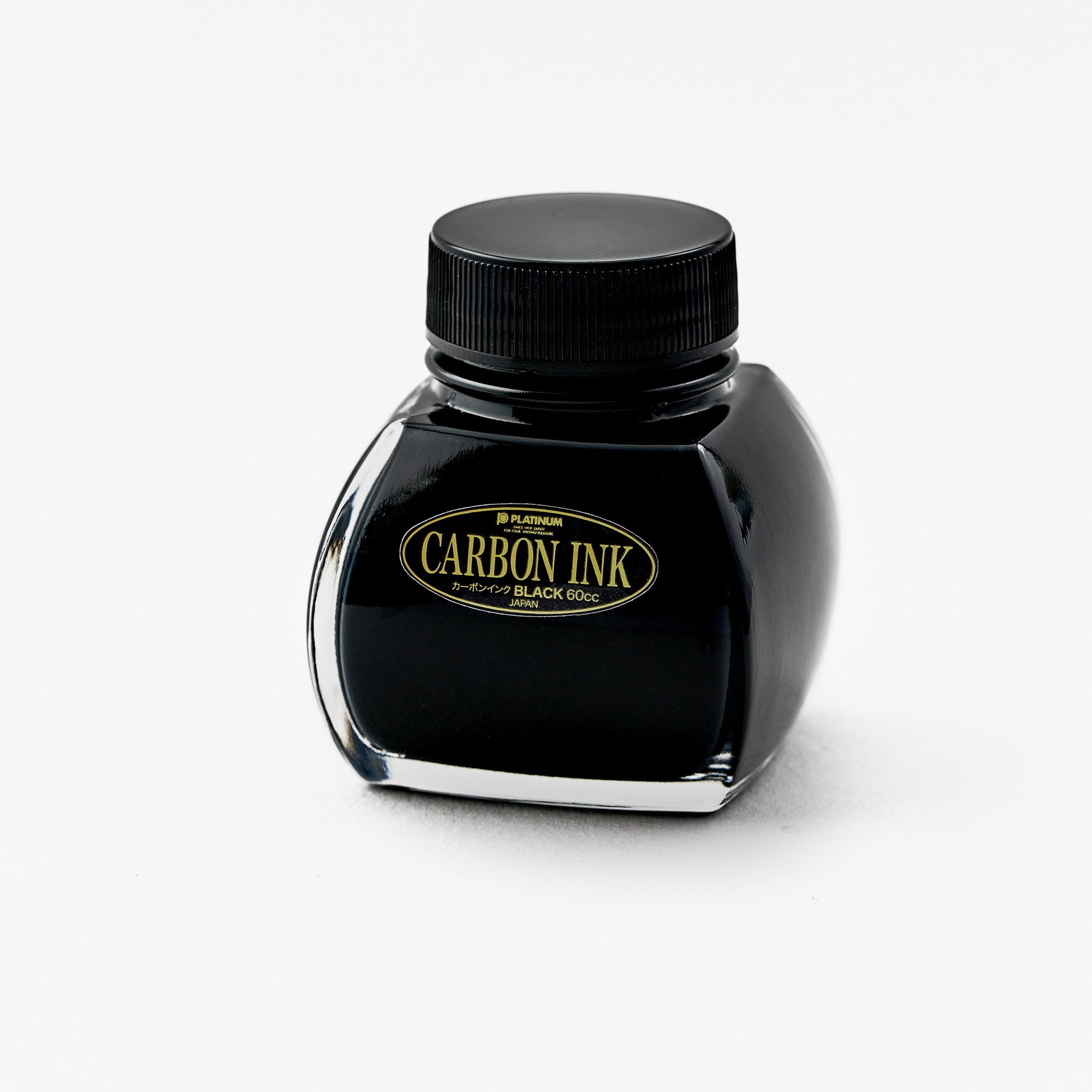 A bottle of Platinum Carbon Ink is positioned at a slight angle, showing the left side of the bottle. The ink is housed in a uniquely shaped glass bottle with a black screw-on lid. The label is gold and black, featuring the words "Carbon Ink" prominently in the centre.