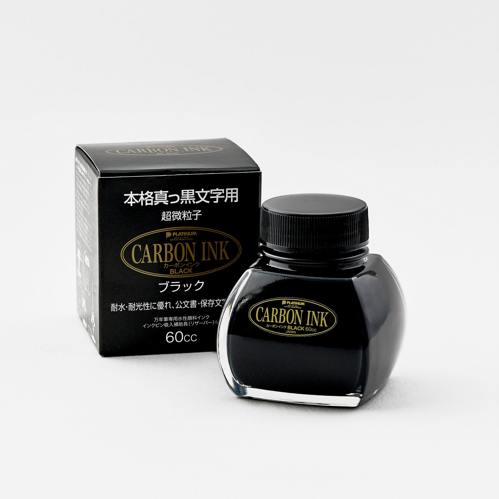 A Platinum Carbon Ink bottle placed next to its original packaging. The box is black with gold and white Japanese text, complementing the bottle’s gold and black label. The bottle's signature curved shape is visible, along with its black screw cap.