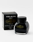 A Platinum Carbon Ink bottle placed next to its original packaging. The box is black with gold and white Japanese text, complementing the bottle’s gold and black label. The bottle's signature curved shape is visible, along with its black screw cap.