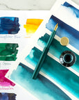 The Pocket Fountain Pen in Ivy shown with an open bottle of fountain pen inks and a selection of ink colour swatches on paper