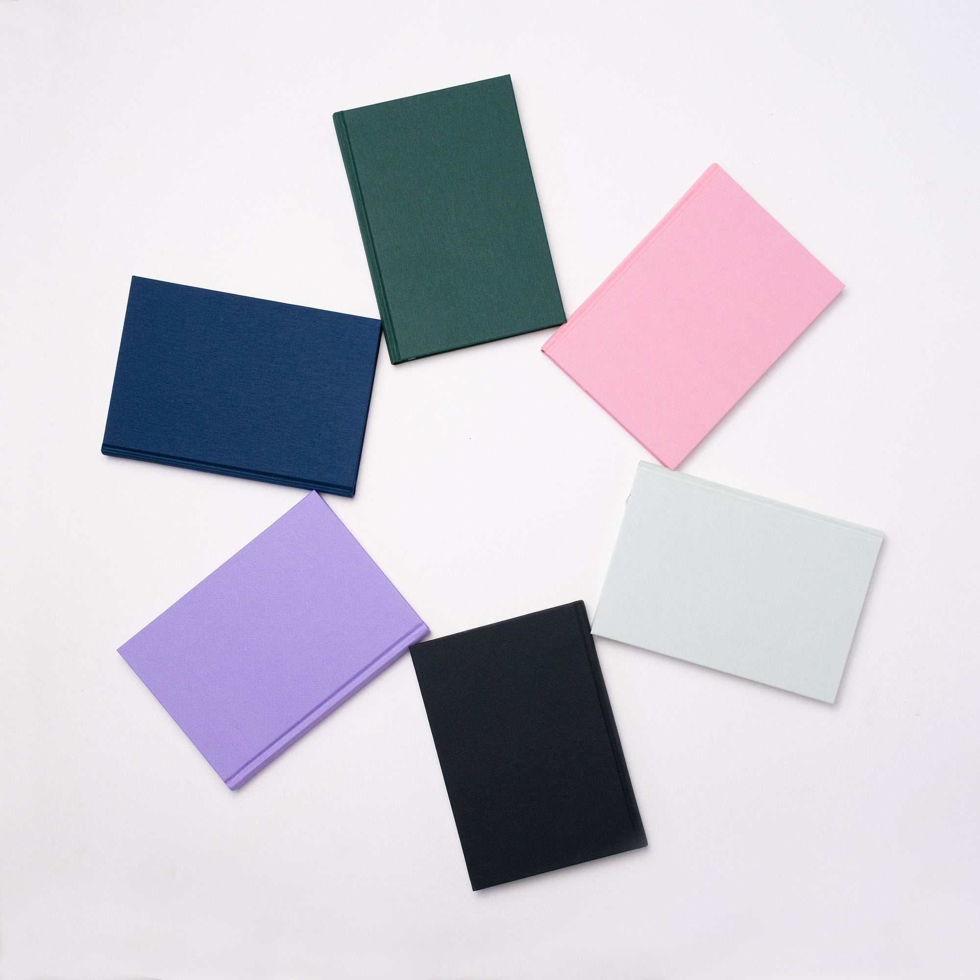 Six Semikolon A5 notebooks in a circular arrangement, featuring vibrant colours like Forest green, Flamingo pink, Moss green, Lilac, Black and Marine blue, with premium linen bound covers.