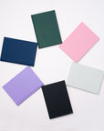 Six Semikolon A5 notebooks in a circular arrangement, featuring vibrant colours like Forest green, Flamingo pink, Moss green, Lilac, Black and Marine blue, with premium linen bound covers.