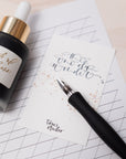 A straight nib calligraphy pen shown with writing on a notepad and spare ink