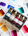 Four bottles of Tom's Studio Calligraphy Ink packaged in a gift box surrounded by ink swatches showing all available colours