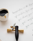 The Flourish calligraphy pen with oblique nib setting in Black on a brass pen rest on top of a sheet of modern calligraphy with an ink well
