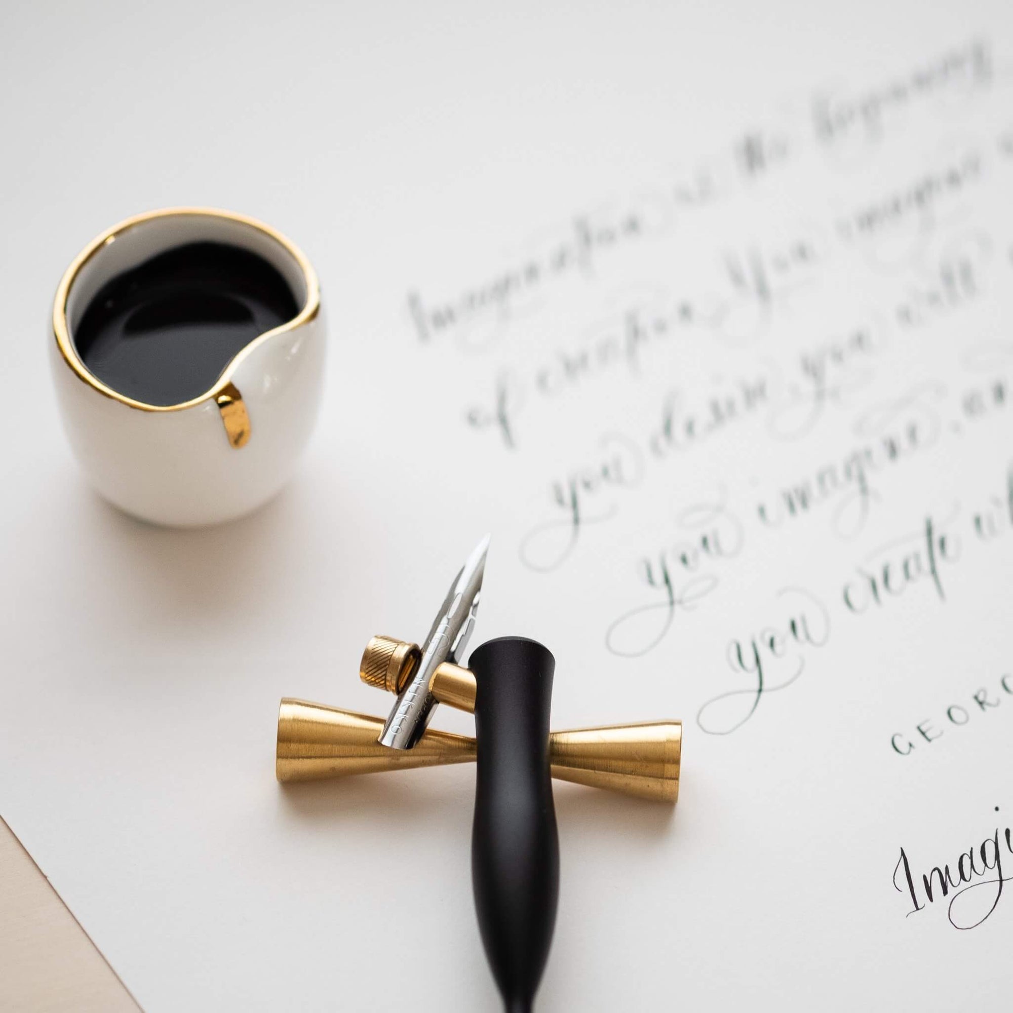 black oblique calligraphy pen on a brass pen rest resting on a sheet of modern calligraphy with an ink well