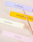 A scented pencil resting on the prism box packaging with the four different scents on display (top to bottom) Lavender, Orange Blossom, Jasmine and Peony