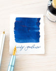 Tom's Studio Royal Blue Fountain Pen Ink with two pens with inky goodness on paper with an ink swatch demonstrating the colour
