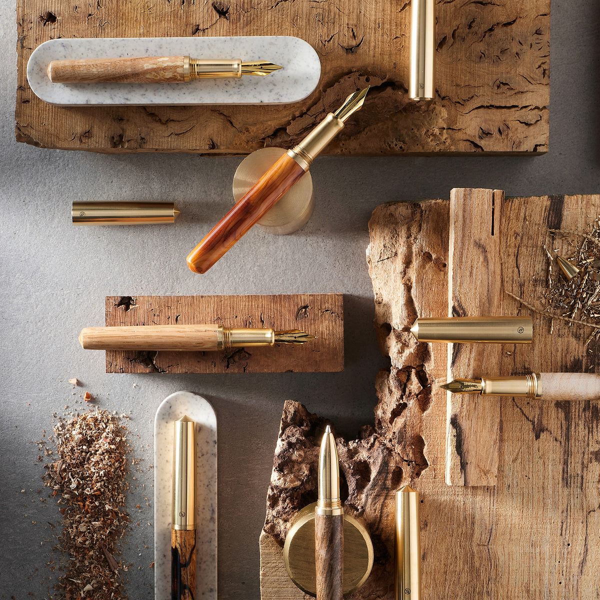 An overhead flat-lay of multiple fountain pens and pen components arranged on rustic wood blocks and boards against a dark gray surface. The pens feature wood-grain handles, brass metal bodies, and some with white or cream grips.