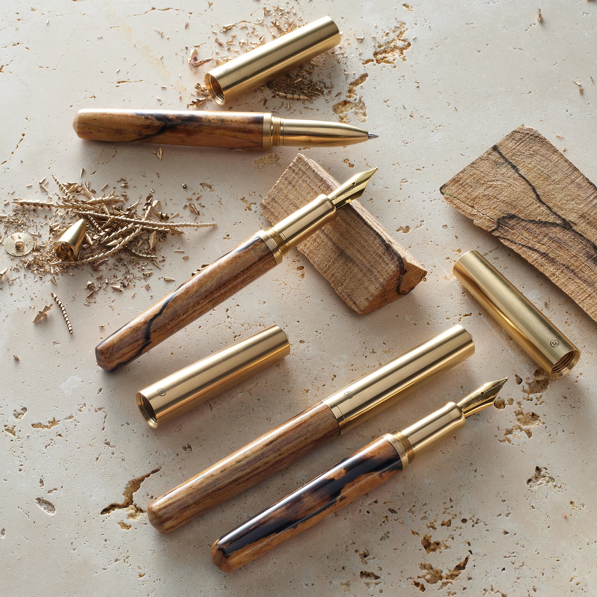 Four wooden fountain pens with gold-toned metal nibs and caps are arranged on a light textured surface with wood shavings and small wood pieces. No person, animal, or real environment is present, so this is Product/Packaging rather than Lifestyle.