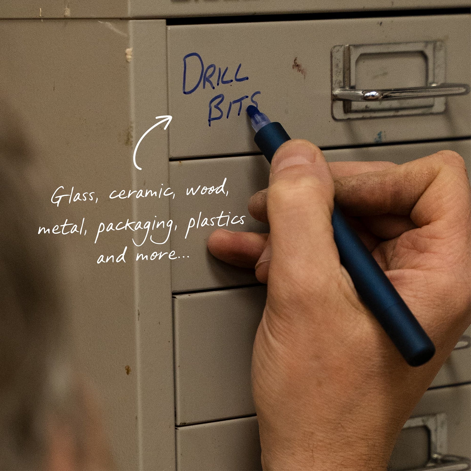 A close-up of a hand holding a dark blue marker and writing on a beige metal cabinet door. White handwritten text on the left reads 'DRILL BITS' with an arrow pointing to the writing, and additional text lists materials such as glass, ceramic, wood, metal, packaging, and plastics.