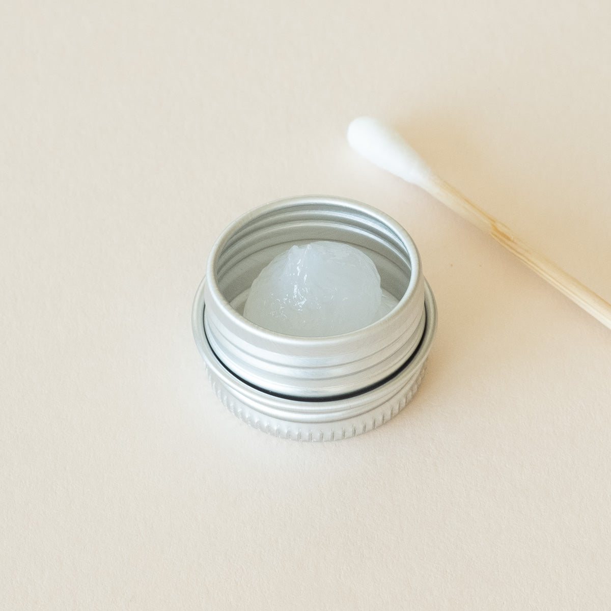 An open metal tin containing silicone grease, with a cotton bud placed beside it, displayed on a light beige background.