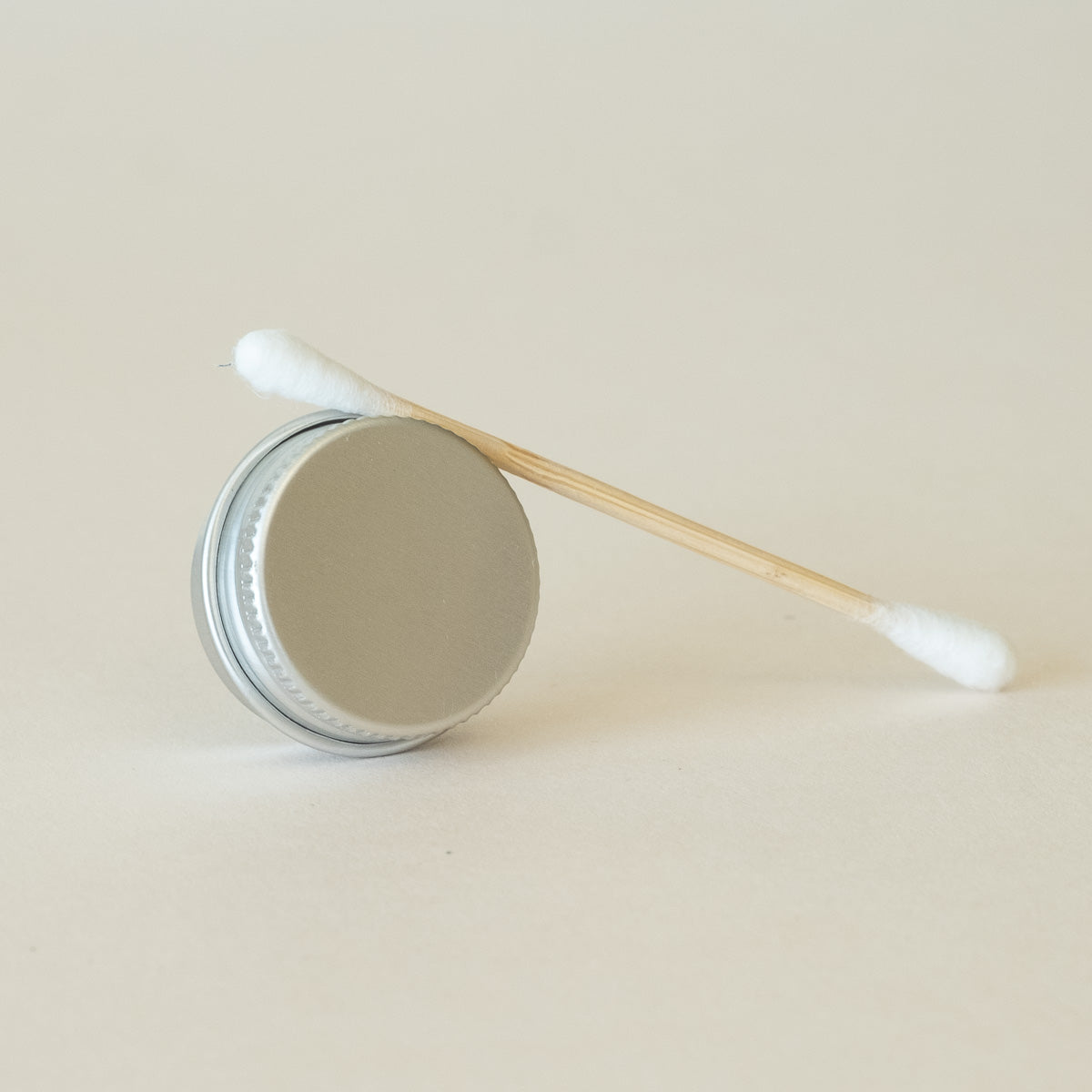 A closed metal circular tin positioned on its side with a cotton bud resting on top, displayed on a light beige background.