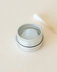 An open metal tin containing silicone grease, with a cotton bud placed beside it, displayed on a light beige background.