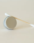 A closed metal circular tin positioned on its side with a cotton bud resting on top, displayed on a light beige background.