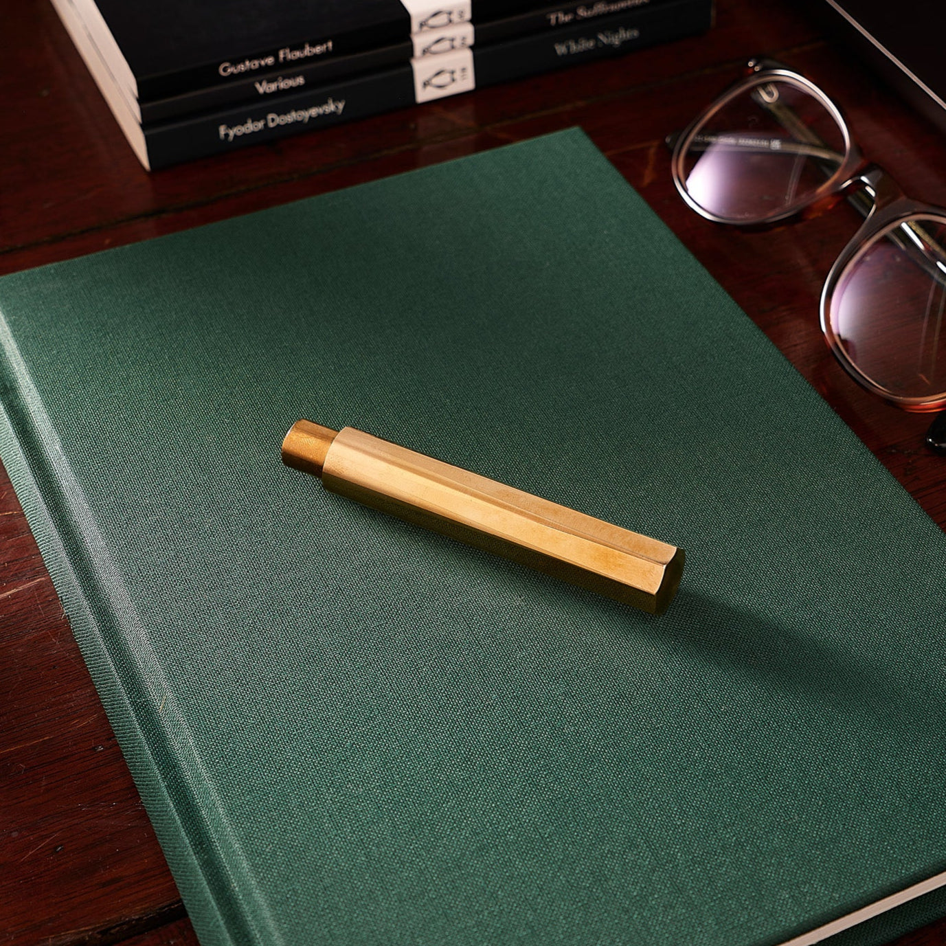 A Brass Wren refillable writing pen , capped, rests on top of a dark green notebook, placed beside glasses, a laptop, and stacked paperbacks on a wooden desk.