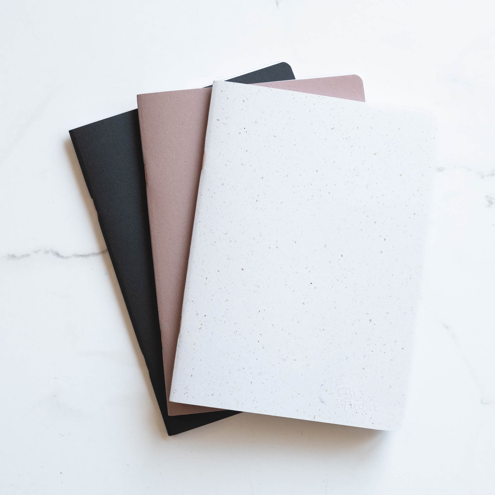 Three A5 Tom's Studio x Coffeenotes Notebooks in Black, Almond, and 'Grounds' (white), made from recycled coffee cups and eco-friendly materials.