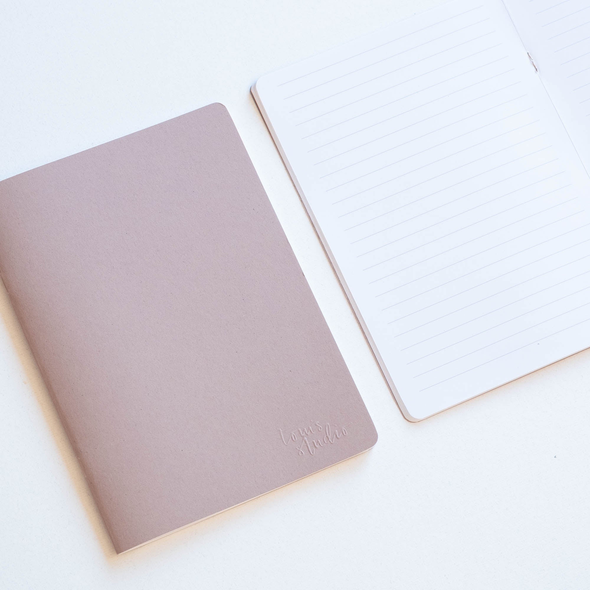 Two Tom's Studio A5 coffee cup notebooks sitting side by side on a table, a almond coloured notebook is shut and the other open.