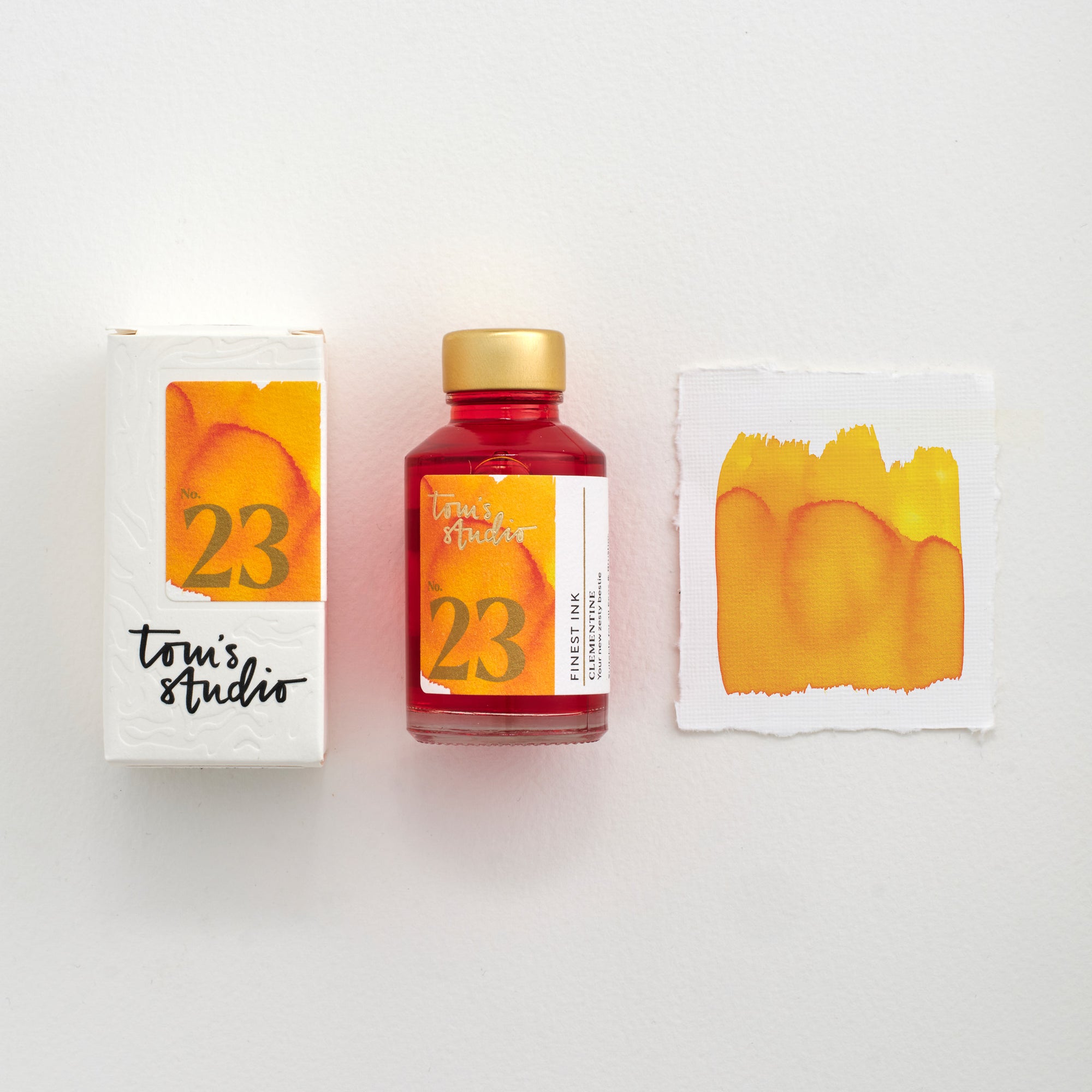 Tom's Studio Clementine Fountain Pen Ink – bottle with gold lid, boxed packaging featuring an orange and gold label marked '23', and a swatch of the vibrant orange ink on textured paper.