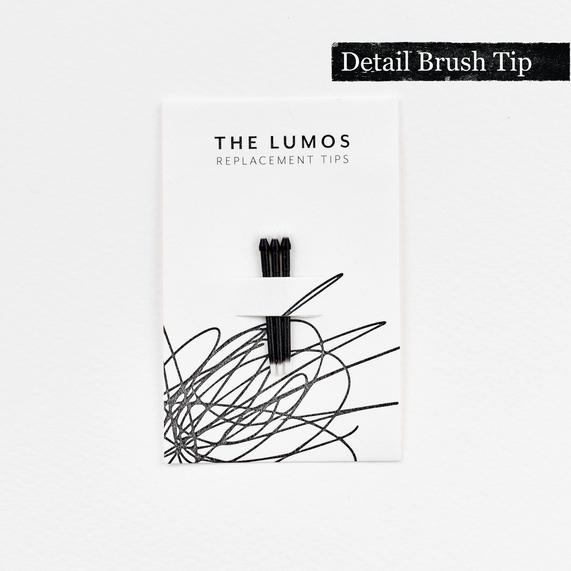 A pack of three Lumos replacement Detail Brush tips attached to a white backing card. The card features 'THE LUMOS REPLACEMENT TIPS' in uppercase black letters at the top, with an abstract black ink scribble design in the lower half. A black textured label in the upper right corner displays 'Detail Brush Tip' in white text. 