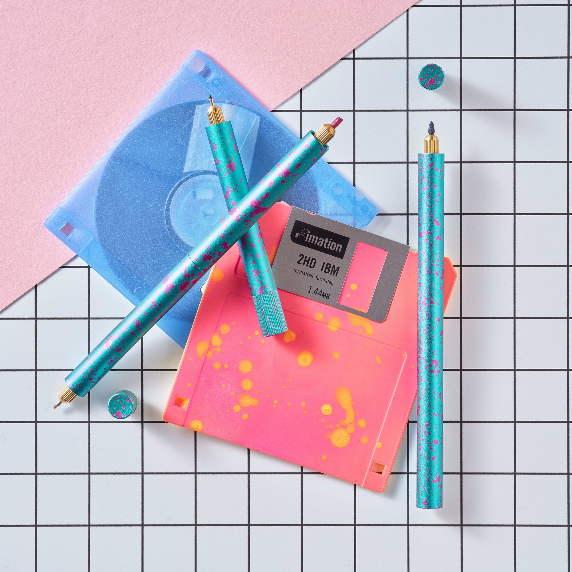 Close-up of Lumos Splash pens in Blue Raspberry colourway with pink splatter, styled with neon pink and blue floppy disks on a grid background.