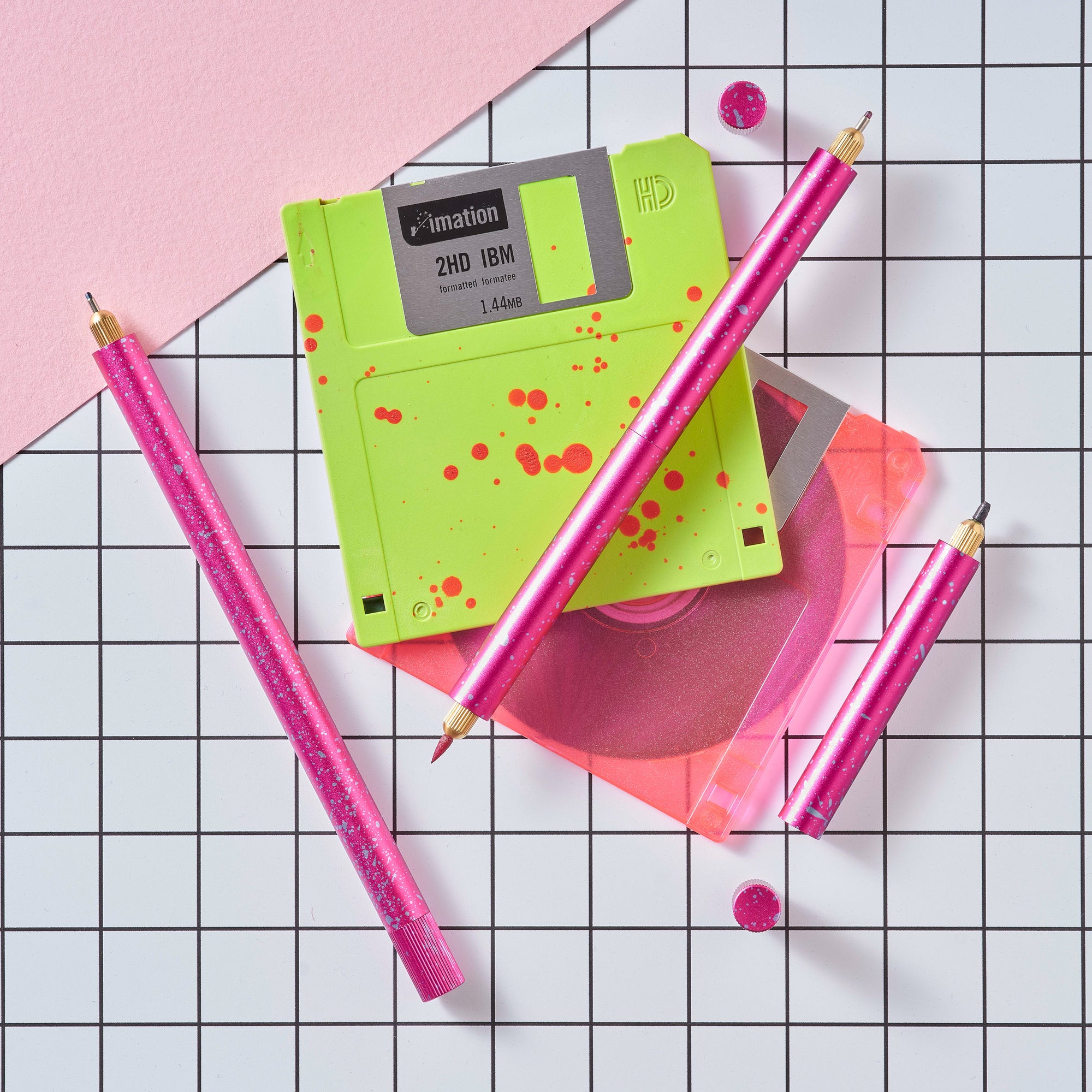 Close-up of Lumos Splash pens in Bubblegum pink with blue splatter, styled with neon green and pink floppy disks on a grid background.