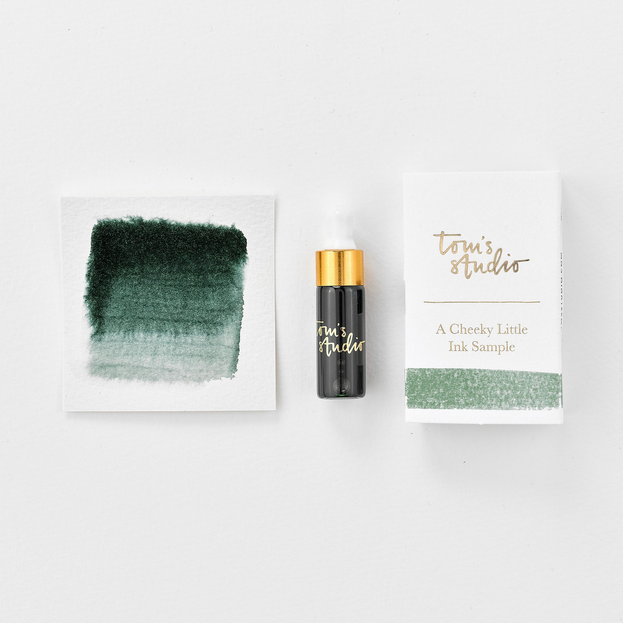 A mini ink sample bottle of Ivy green fountain pen ink by Tom’s Studio, displayed alongside a swatch of the ink on textured paper and its matching packaging. The bottle is small, with a gold cap and a white pipette dropper, and features 'Tom’s Studio' in gold lettering. The ink swatch shows a rich, shimmering hue with a gradient from deep to lighter tones. The packaging, a small white card, has 'Tom’s Studio' embossed in gold at the top, with a brushstroke of the ink colour at the bottom.