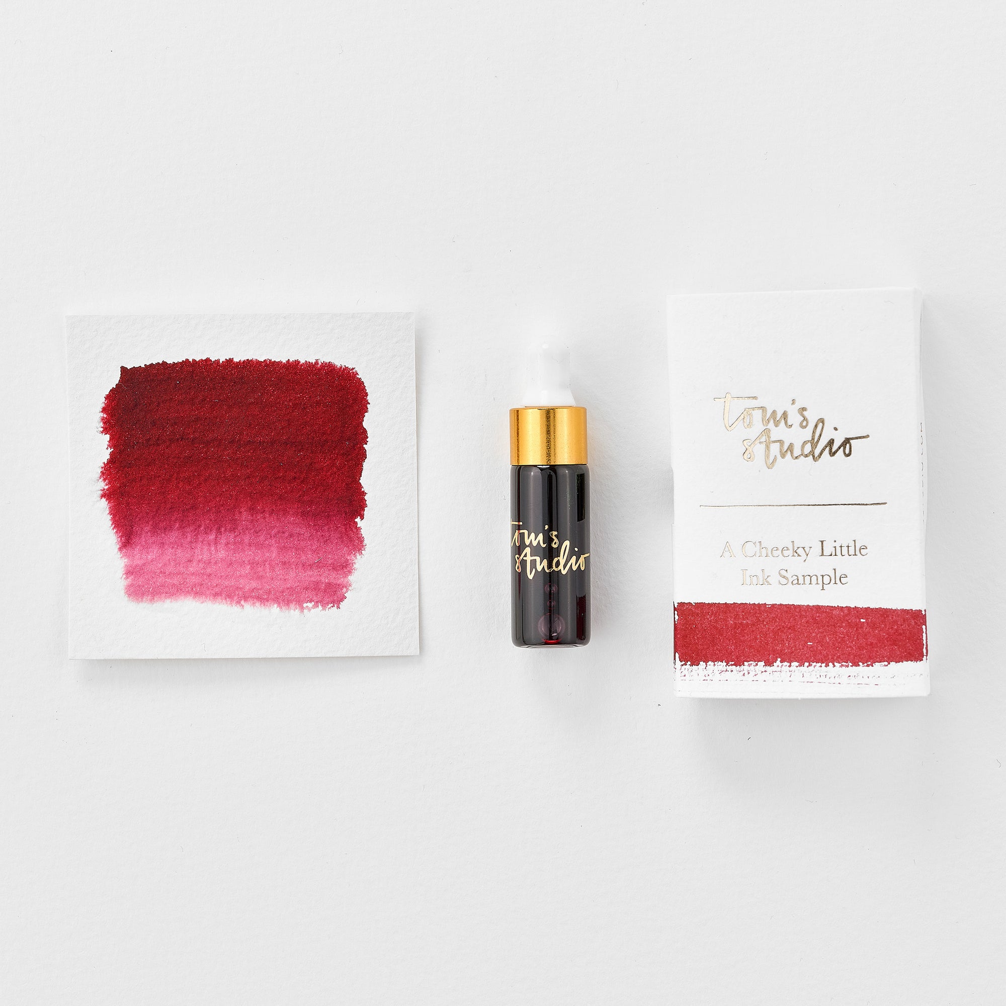 A mini ink sample bottle of Cassis red fountain pen ink by Tom’s Studio, displayed alongside a swatch of the ink on textured paper and its matching packaging. The bottle is small, with a gold cap and a white pipette dropper, and features 'Tom’s Studio' in gold lettering. The ink swatch shows a rich, shimmering hue with a gradient from deep to lighter tones. The packaging, a small white card, has 'Tom’s Studio' embossed in gold at the top, with a brushstroke of the ink colour at the bottom.