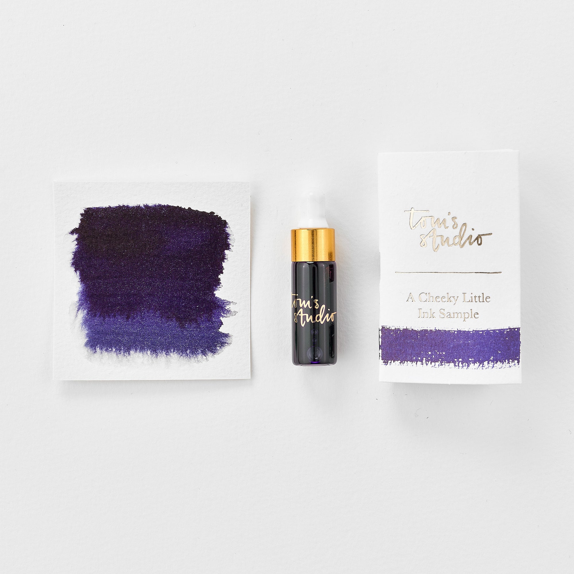 A mini ink sample bottle of Constellation purple fountain pen ink by Tom’s Studio, displayed alongside a swatch of the ink on textured paper and its matching packaging. The bottle is small, with a gold cap and a white pipette dropper, and features &#39;Tom’s Studio&#39; in gold lettering. The ink swatch shows a rich, shimmering hue with a gradient from deep to lighter tones. The packaging, a small white card, has &#39;Tom’s Studio&#39; embossed in gold at the top, with a brushstroke of the ink colour at the bottom.