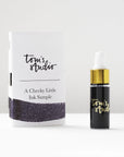 A mini ink sample bottle of Constellation purple fountain pen ink by Tom’s Studio, displayed alongside a swatch of the ink on textured paper and its matching packaging. The bottle is small, with a gold cap and a white pipette dropper, and features 'Tom’s Studio' in gold lettering. The ink swatch shows a rich, shimmering hue with a gradient from deep to lighter tones. The packaging, a small white card, has 'Tom’s Studio' embossed in gold at the top, with a brushstroke of the ink colour at the bottom.