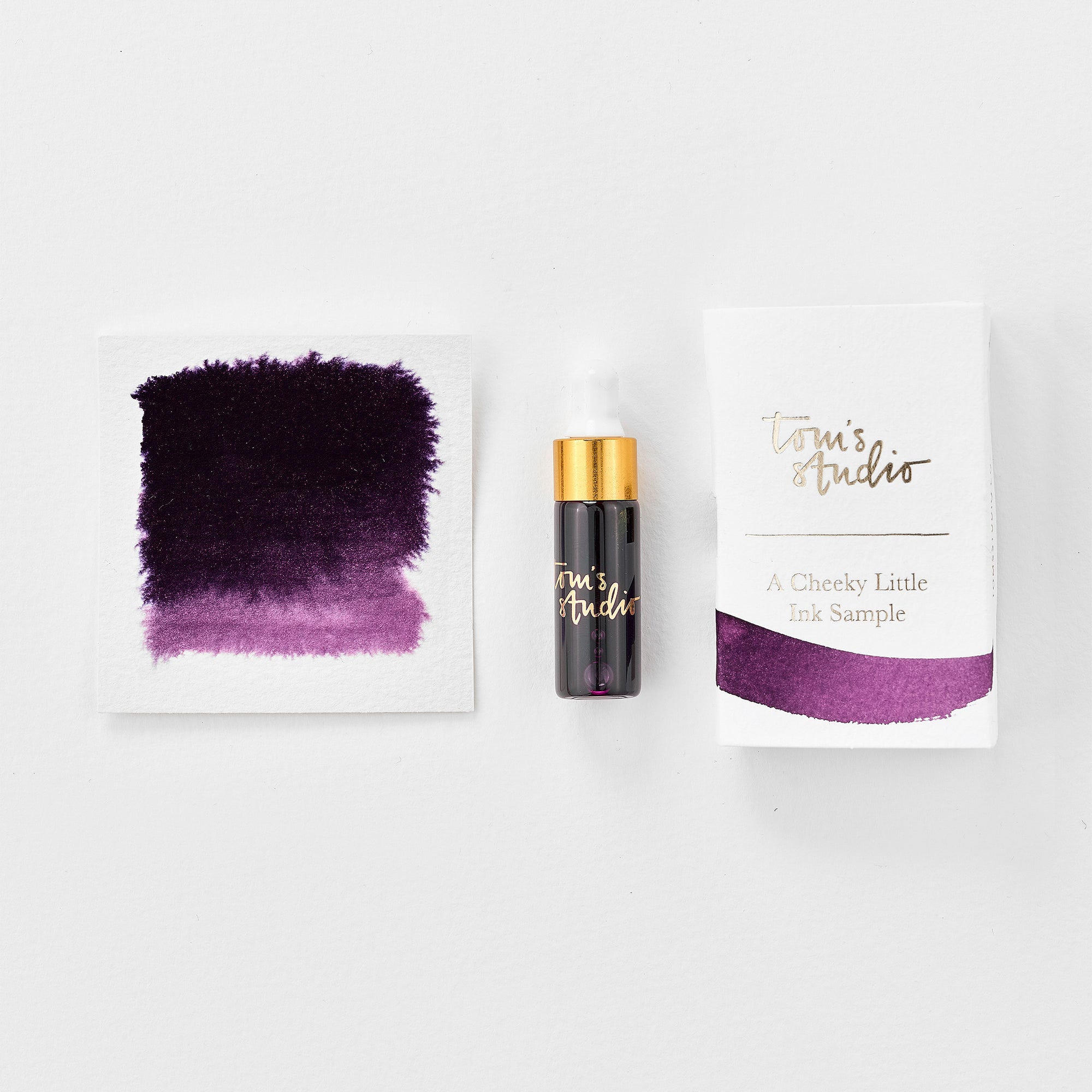 A mini ink sample bottle of Damson purple fountain pen ink by Tom’s Studio, displayed alongside a swatch of the ink on textured paper and its matching packaging. The bottle is small, with a gold cap and a white pipette dropper, and features 'Tom’s Studio' in gold lettering. The ink swatch shows a rich, shimmering hue with a gradient from deep to lighter tones. The packaging, a small white card, has 'Tom’s Studio' embossed in gold at the top, with a brushstroke of the ink colour at the bottom.