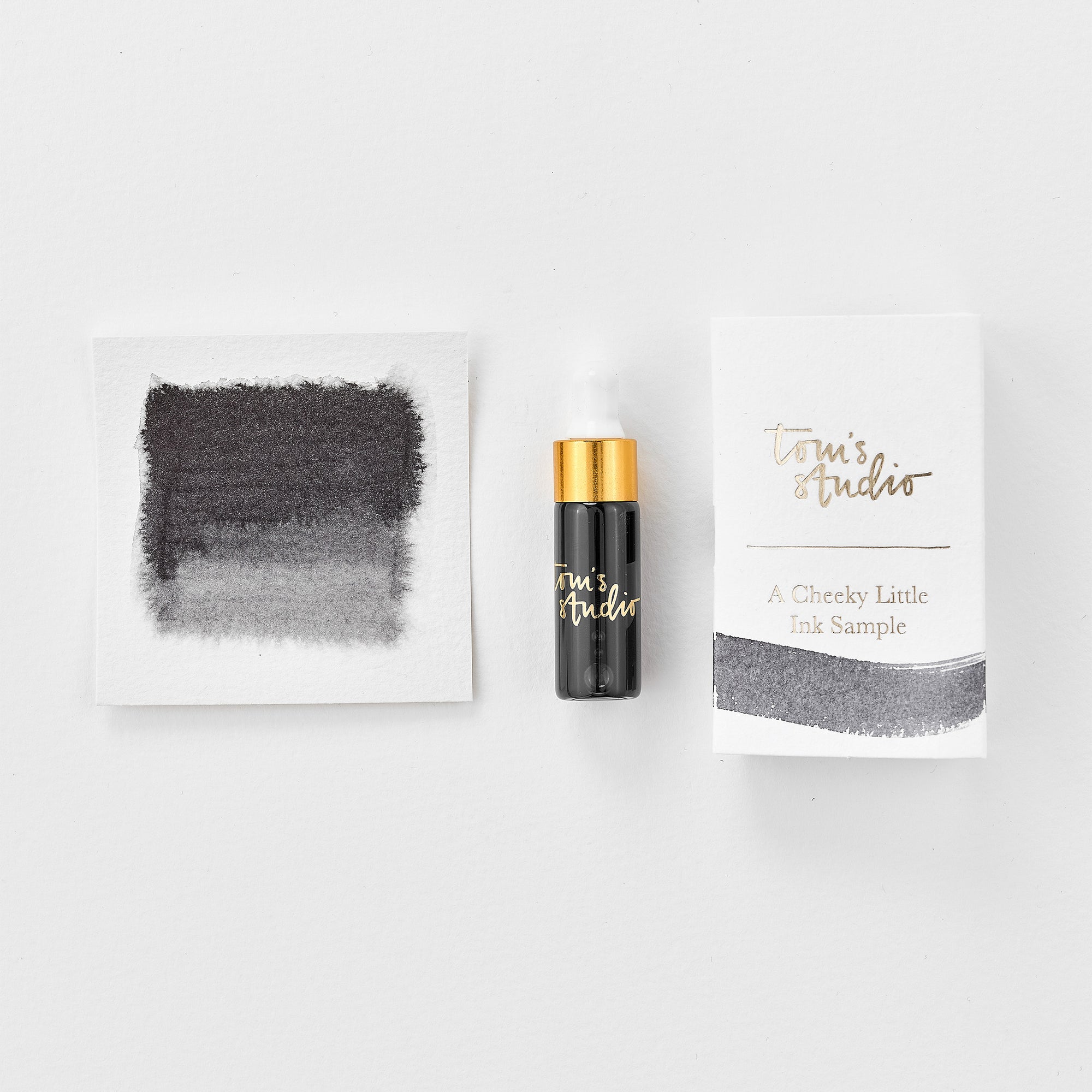 A mini ink sample bottle of Dove Grey fountain pen ink by Tom’s Studio, displayed alongside a swatch of the ink on textured paper and its matching packaging. The bottle is small, with a gold cap and a white pipette dropper, and features 'Tom’s Studio' in gold lettering. The ink swatch shows a rich, shimmering hue with a gradient from deep to lighter tones. The packaging, a small white card, has 'Tom’s Studio' embossed in gold at the top, with a brushstroke of the ink colour at the bottom.