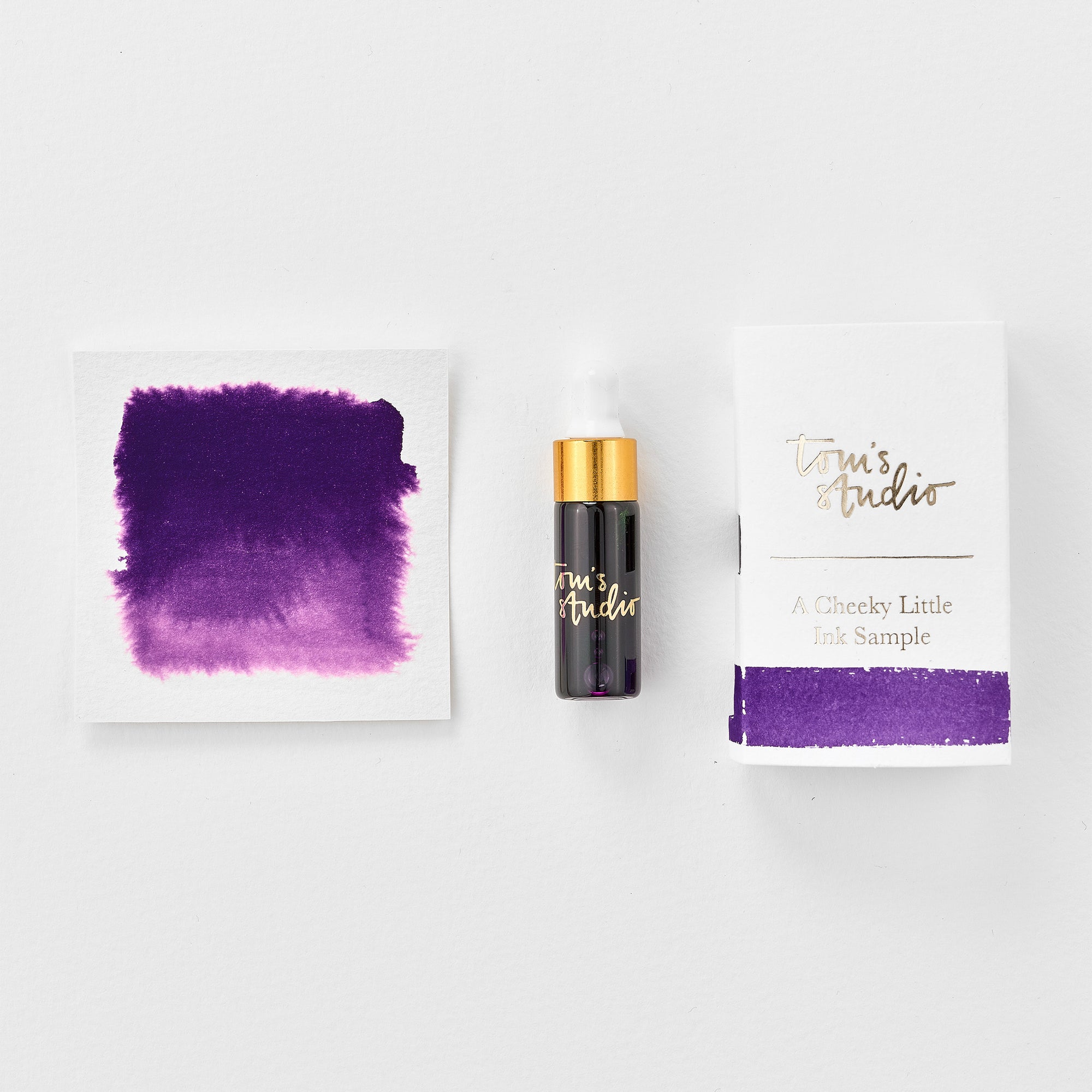 A mini ink sample bottle of Iris purple fountain pen ink by Tom’s Studio, displayed alongside a swatch of the ink on textured paper and its matching packaging. The bottle is small, with a gold cap and a white pipette dropper, and features 'Tom’s Studio' in gold lettering. The ink swatch shows a rich, shimmering hue with a gradient from deep to lighter tones. The packaging, a small white card, has 'Tom’s Studio' embossed in gold at the top, with a brushstroke of the ink colour at the bottom.