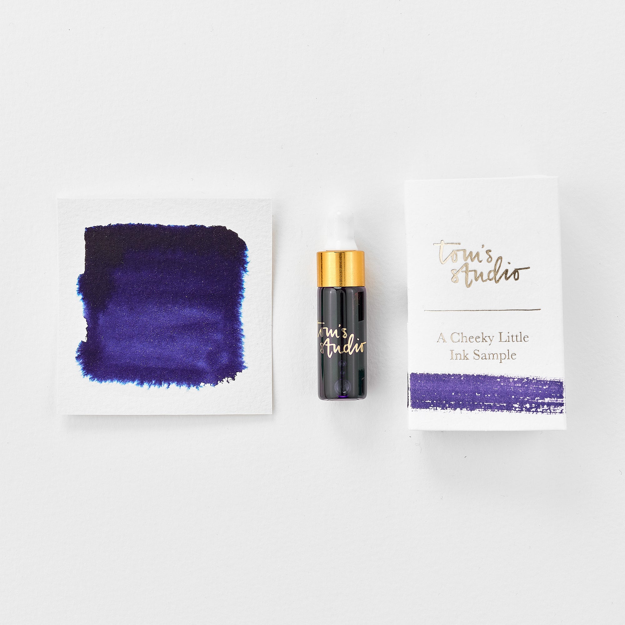 A mini ink sample bottle of Juniper purple fountain pen ink by Tom’s Studio, displayed alongside a swatch of the ink on textured paper and its matching packaging. The bottle is small, with a gold cap and a white pipette dropper, and features 'Tom’s Studio' in gold lettering. The ink swatch shows a rich, shimmering hue with a gradient from deep to lighter tones. The packaging, a small white card, has 'Tom’s Studio' embossed in gold at the top, with a brushstroke of the ink colour at the bottom.