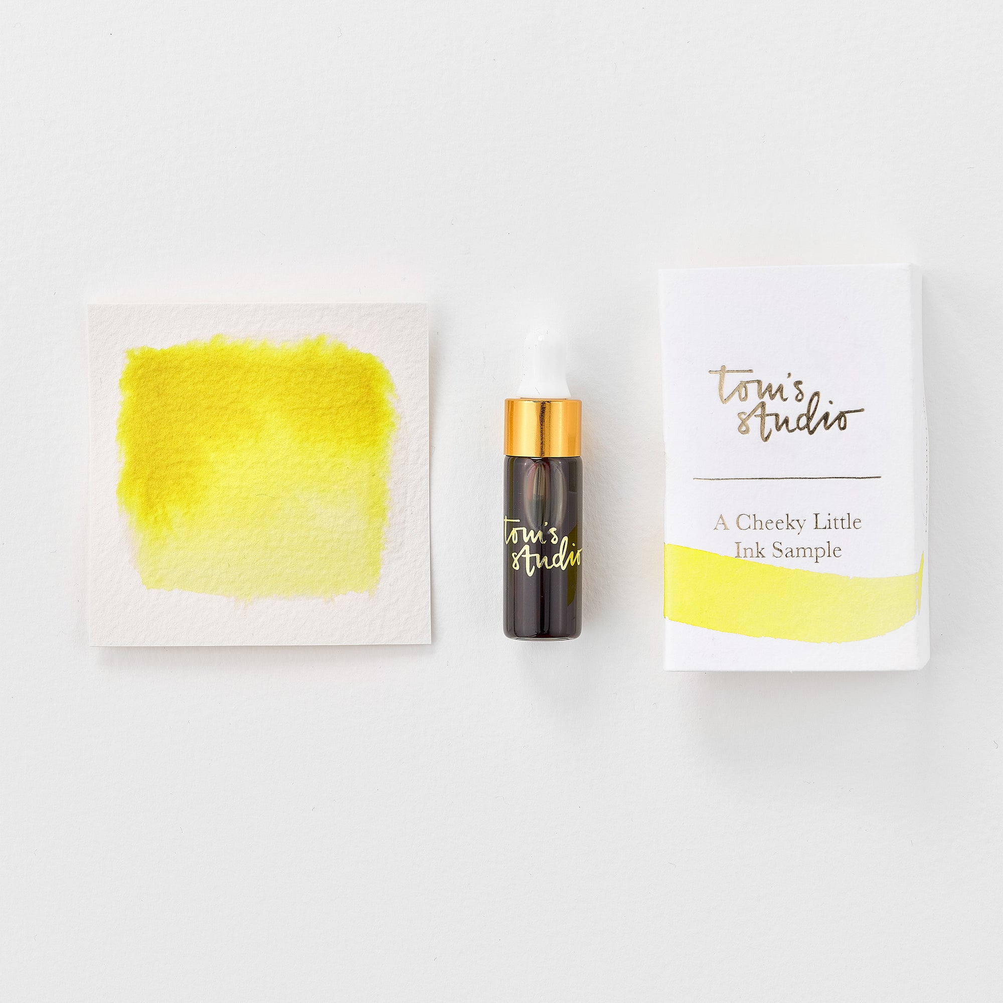 A mini ink sample bottle of Lemon Yellow fountain pen ink by Tom’s Studio, displayed alongside a swatch of the ink on textured paper and its matching packaging. The bottle is small, with a gold cap and a white pipette dropper, and features 'Tom’s Studio' in gold lettering. The ink swatch shows a rich, shimmering hue with a gradient from deep to lighter tones. The packaging, a small white card, has 'Tom’s Studio' embossed in gold at the top, with a brushstroke of the ink colour at the bottom.