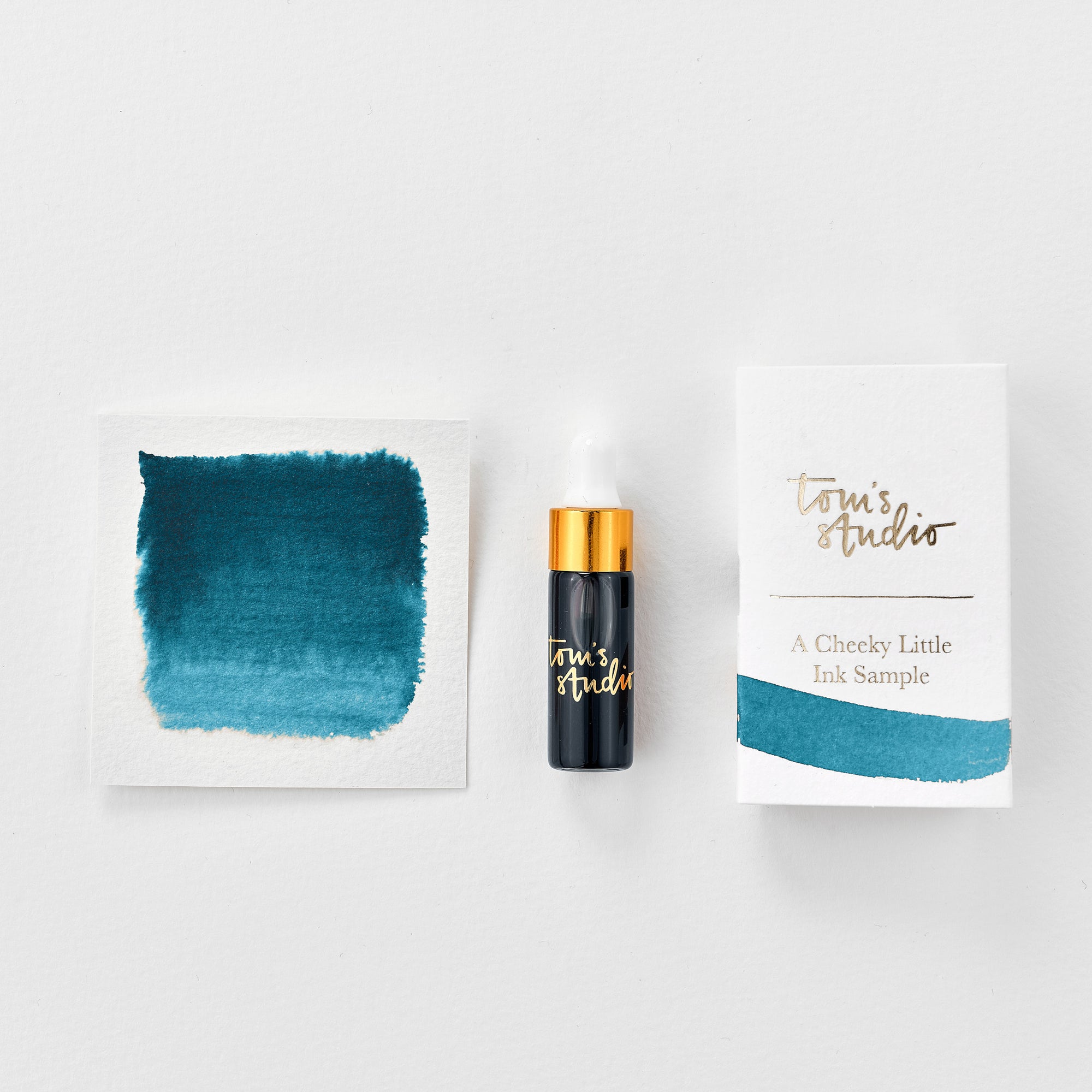 A mini ink sample bottle of Loch Ness blue fountain pen ink by Tom’s Studio, displayed alongside a swatch of the ink on textured paper and its matching packaging. The bottle is small, with a gold cap and a white pipette dropper, and features 'Tom’s Studio' in gold lettering. The ink swatch shows a rich, shimmering hue with a gradient from deep to lighter tones. The packaging, a small white card, has 'Tom’s Studio' embossed in gold at the top, with a brushstroke of the ink colour at the bottom.