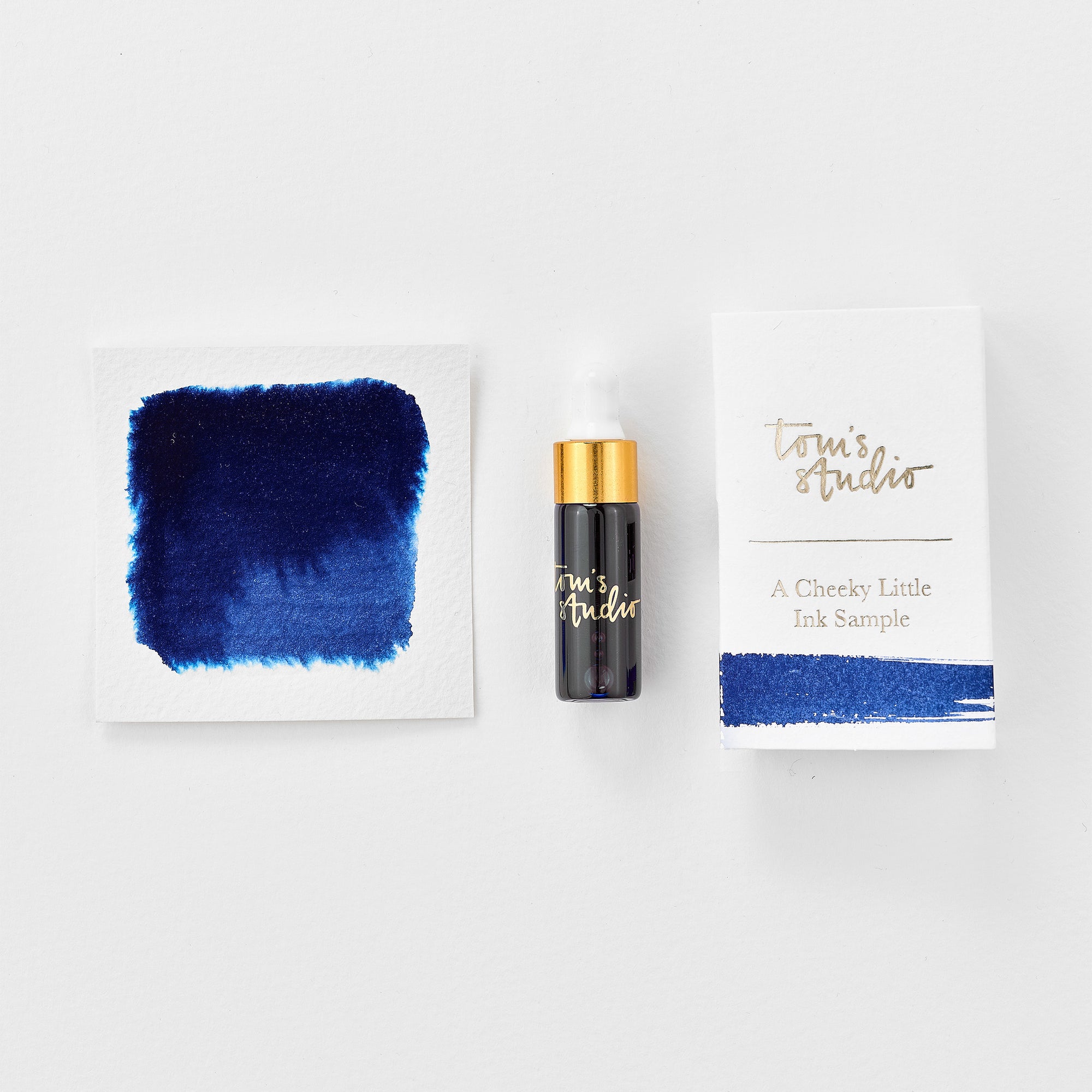 A mini ink sample bottle of Marianas blue fountain pen ink by Tom’s Studio, displayed alongside a swatch of the ink on textured paper and its matching packaging. The bottle is small, with a gold cap and a white pipette dropper, and features 'Tom’s Studio' in gold lettering. The ink swatch shows a rich, shimmering hue with a gradient from deep to lighter tones. The packaging, a small white card, has 'Tom’s Studio' embossed in gold at the top, with a brushstroke of the ink colour at the bottom.