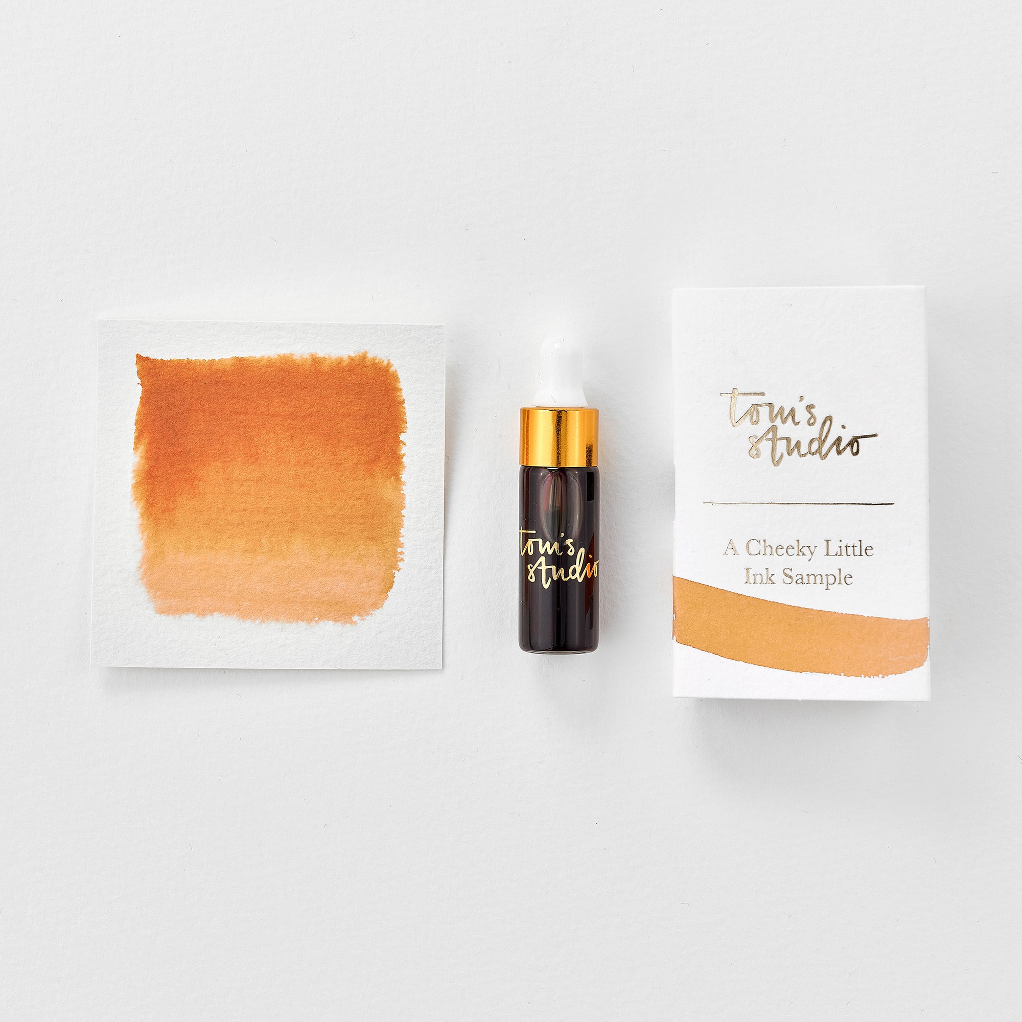 A mini ink sample bottle of Marmalade orange fountain pen ink by Tom’s Studio, displayed alongside a swatch of the ink on textured paper and its matching packaging. The bottle is small, with a gold cap and a white pipette dropper, and features 'Tom’s Studio' in gold lettering. The ink swatch shows a rich, shimmering hue with a gradient from deep to lighter tones. The packaging, a small white card, has 'Tom’s Studio' embossed in gold at the top, with a brushstroke of the ink colour at the bottom.