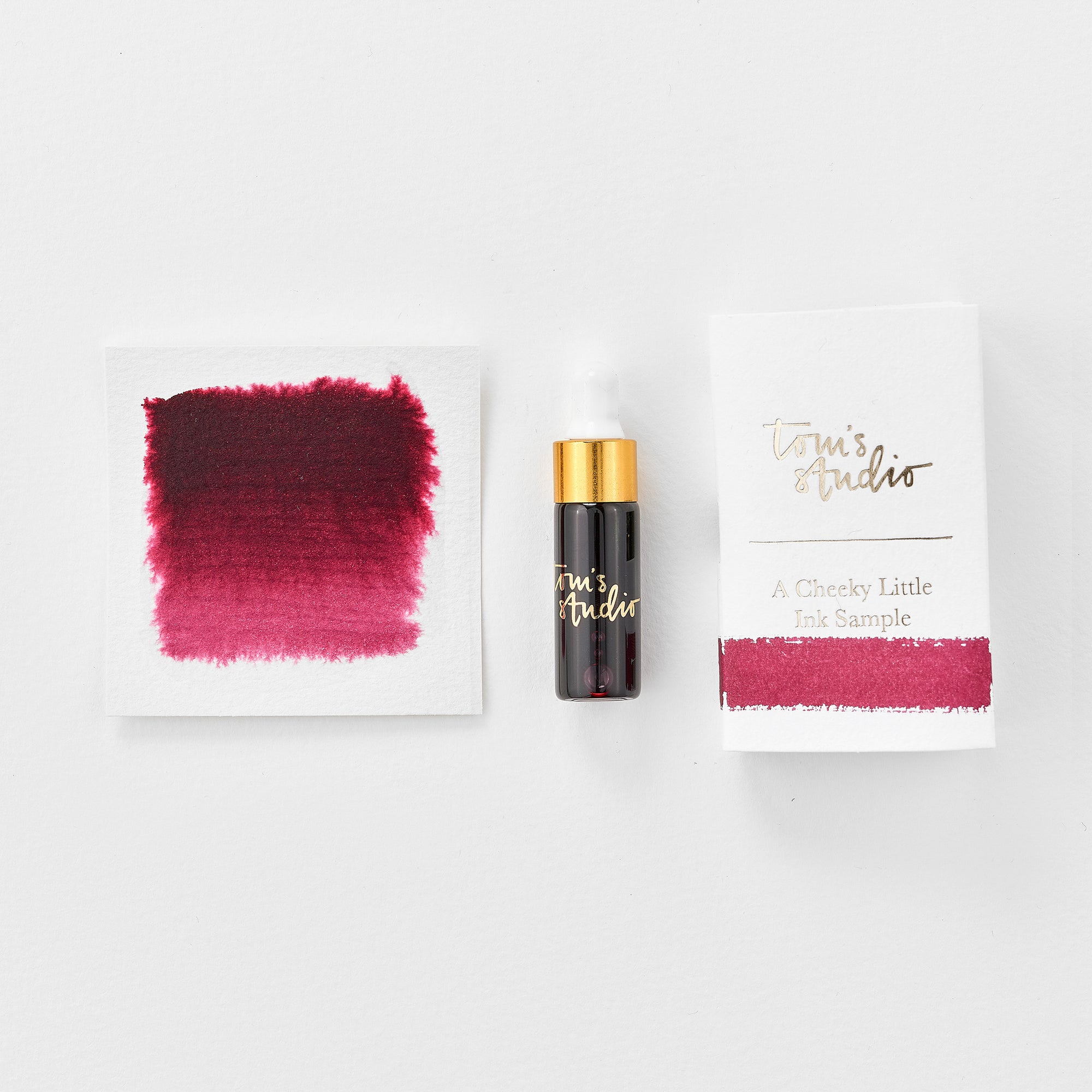 A mini ink sample bottle of Mulberry red fountain pen ink by Tom’s Studio, displayed alongside a swatch of the ink on textured paper and its matching packaging. The bottle is small, with a gold cap and a white pipette dropper, and features 'Tom’s Studio' in gold lettering. The ink swatch shows a rich, shimmering hue with a gradient from deep to lighter tones. The packaging, a small white card, has 'Tom’s Studio' embossed in gold at the top, with a brushstroke of the ink colour at the bottom.